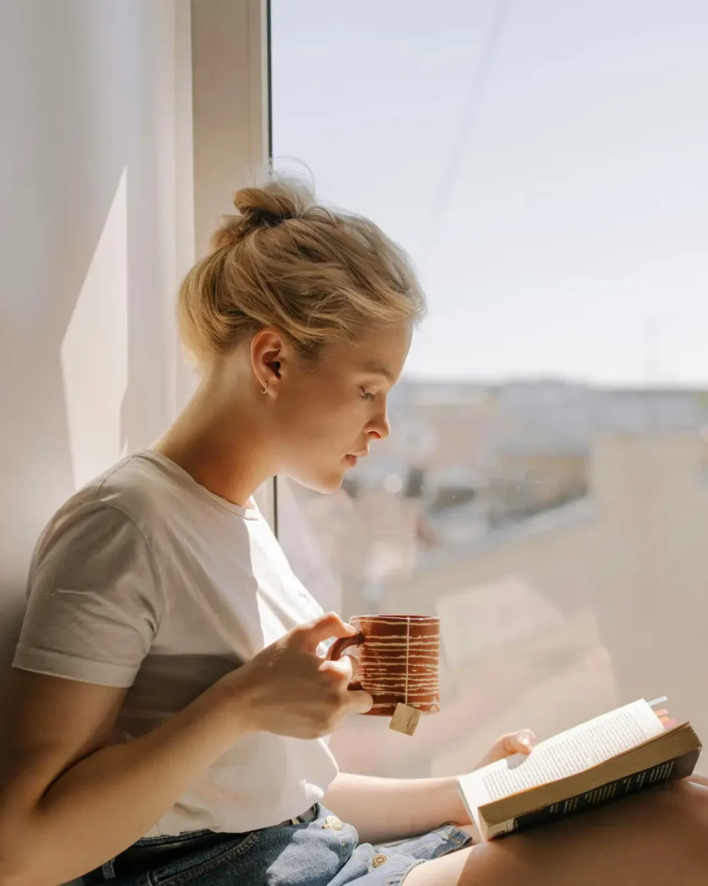 Jeune femme aux cheveux attachés lisant un livre et tenant une tasse de thé, assise au soleil sur le rebord d'une fenêtre.