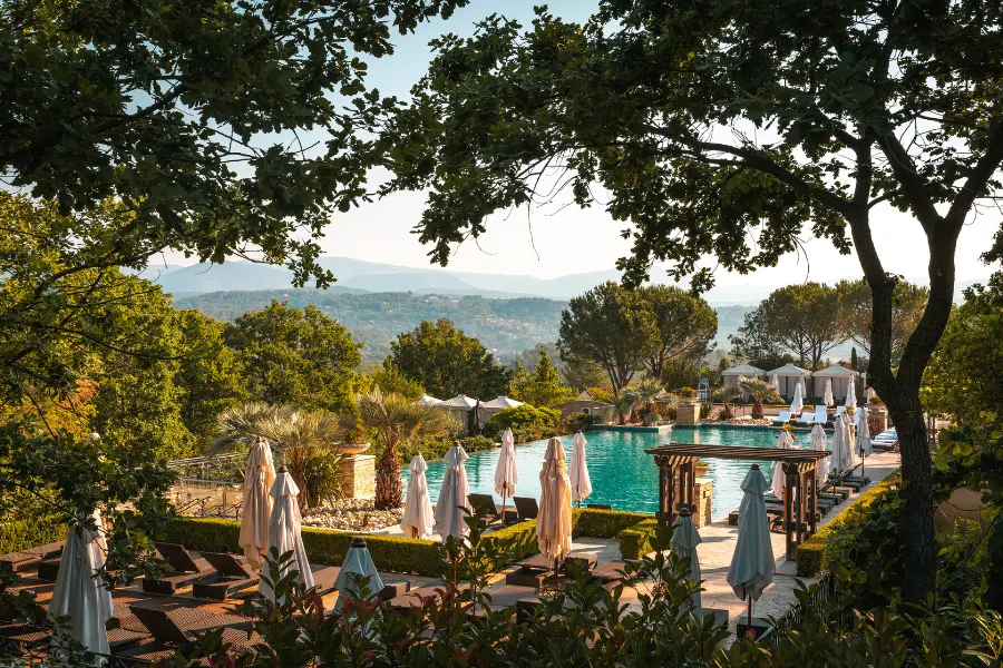 Piscine extérieure à débordement de Terre Blanche entourée de parasols blancs et de transats, avec vue sur les collines de Fayence à travers les arbres.