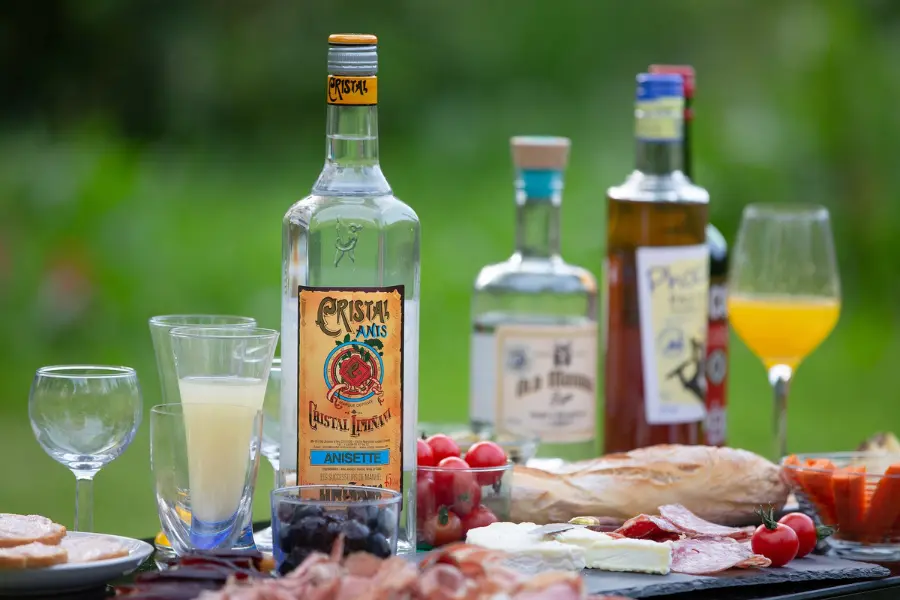 Table d'apéritif avec une bouteille d'anisette Cristal Limiñana, verres de pastis, charcuterie et tomates cerises en extérieur.
